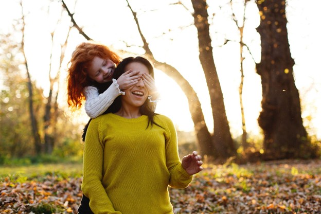 A child covering her mother's eyes while she is kneeling with a fall background with leaves on the ground and trees.